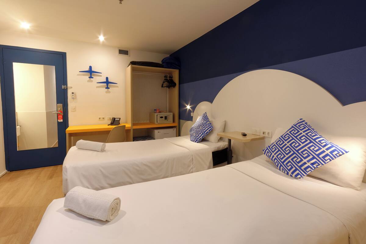 A hotel room with two single beds, a desk, a chair, a mirror, a wardrobe, and a small desk lamp.