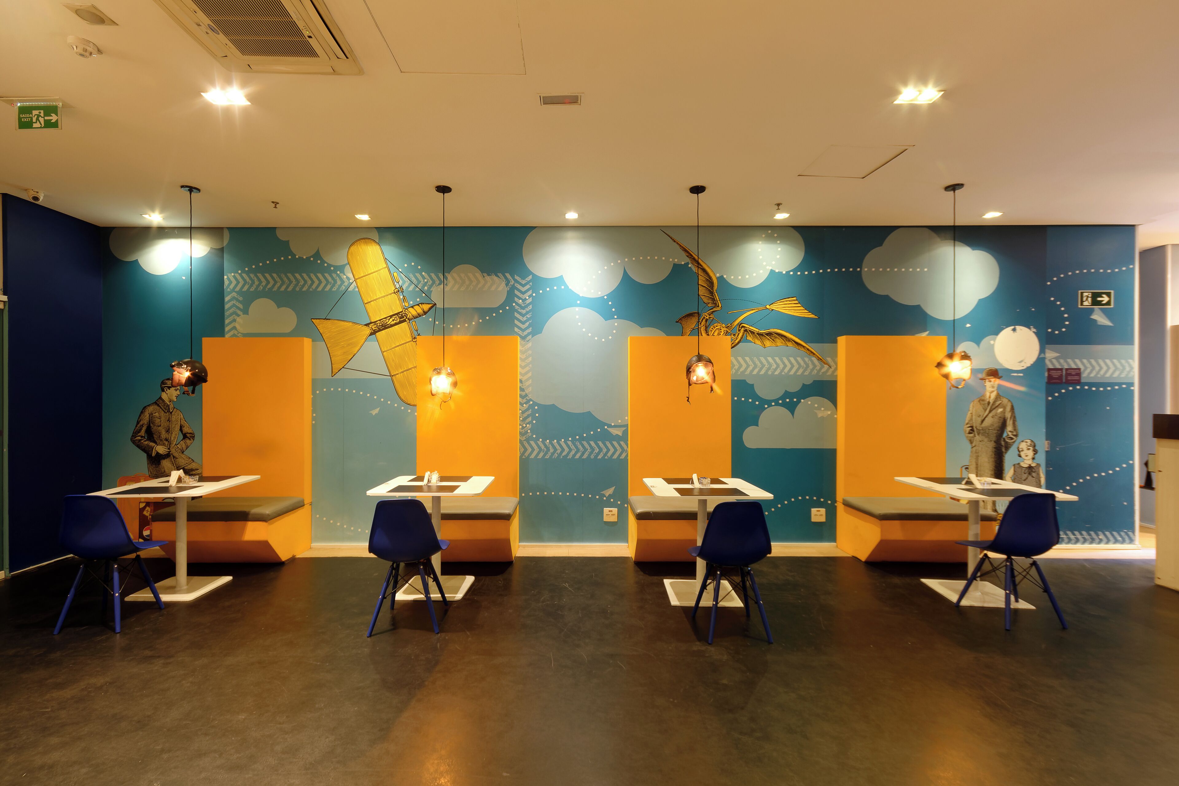 A children's play area with tables and chairs, a mural with clouds and birds, and a blue and orange color scheme.