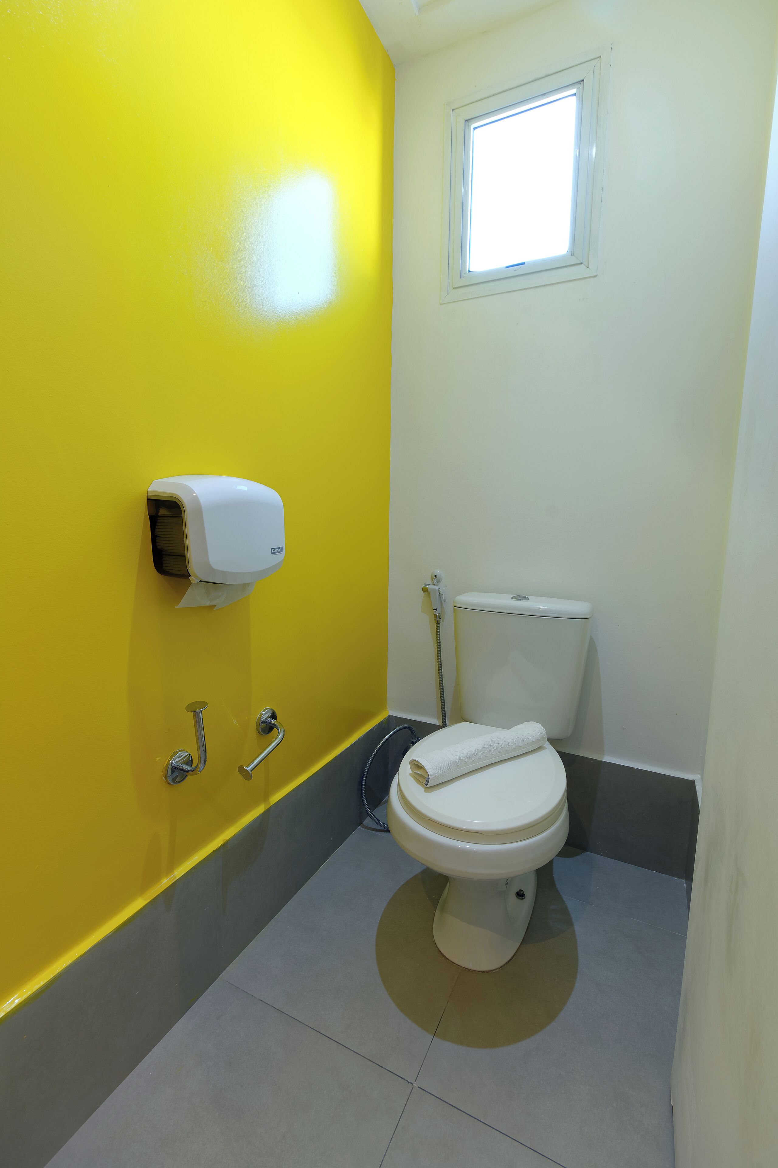 A bathroom with a yellow wall, a white toilet, a hand dryer, and a window.