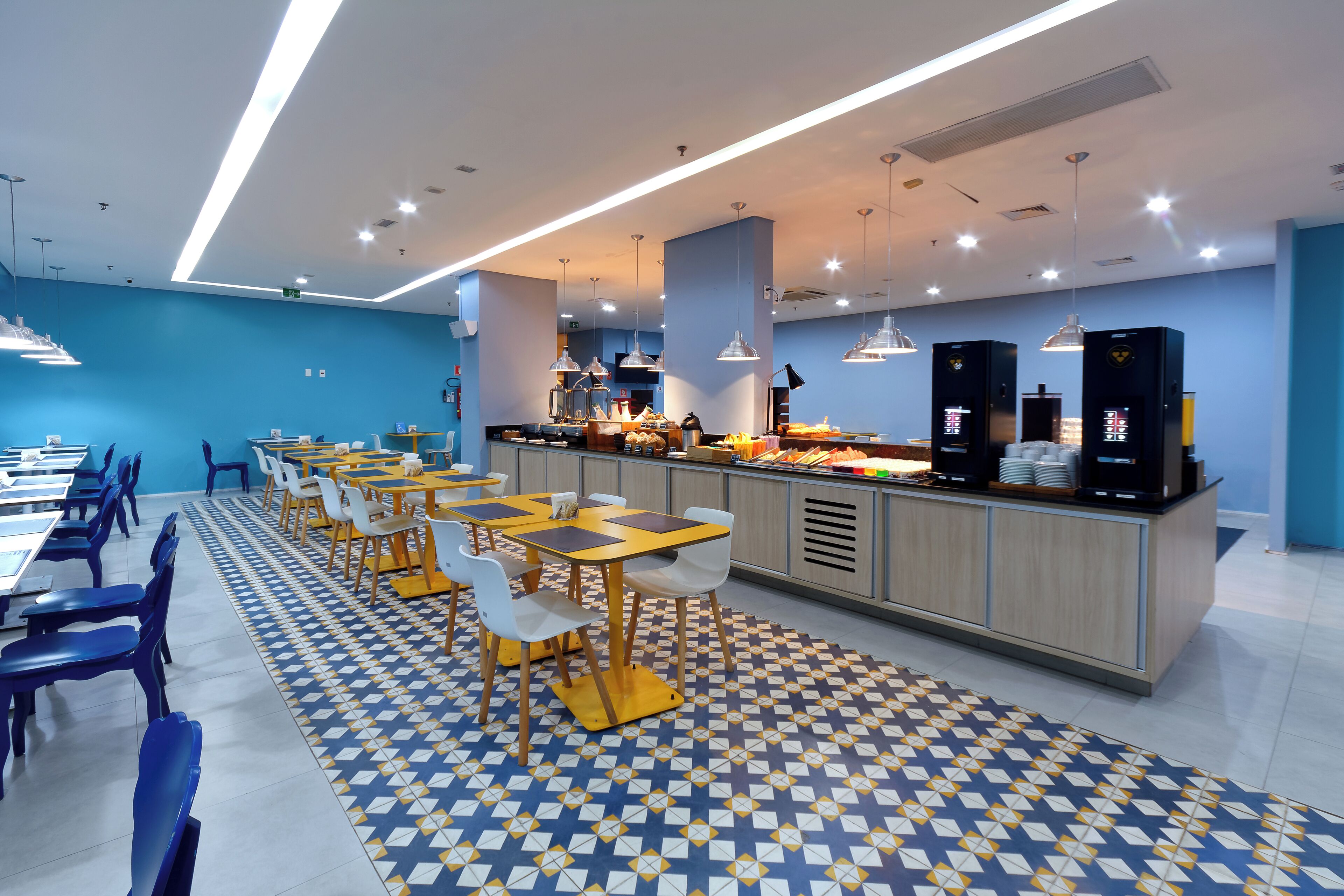A modern cafeteria with multiple tables and chairs, a central counter with food and beverages, and a patterned floor.