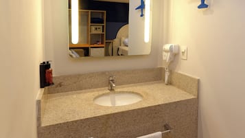 Un baño de hotel con lavamanos de encimera de granito, un espejo con luz, secador de pelo y perchero para toallas.