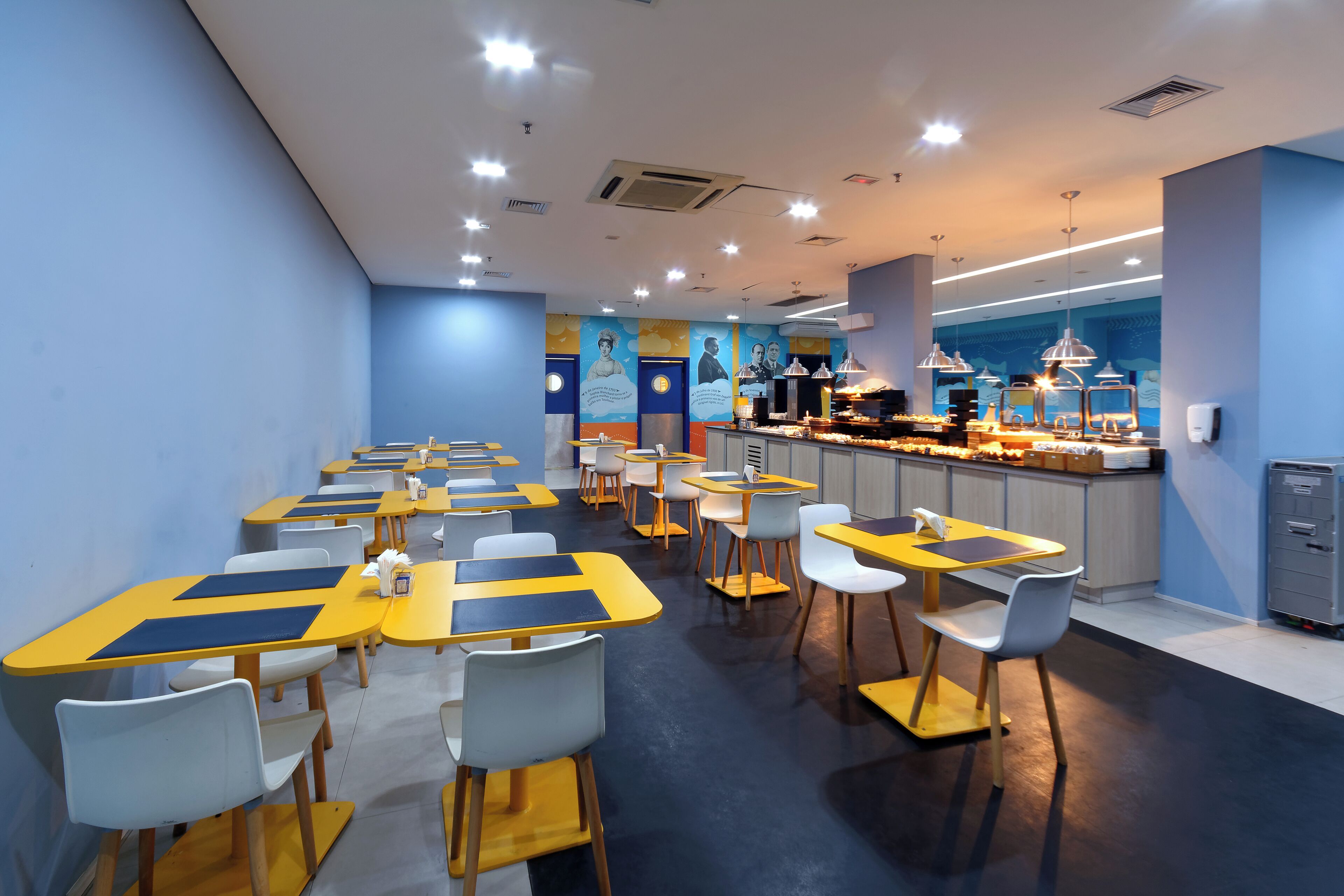A modern restaurant with yellow and blue tables and chairs, a counter with food displays, and a mural on the wall.