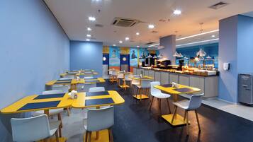 A modern restaurant with yellow and blue tables and chairs, a counter with food displays, and a mural on the wall.