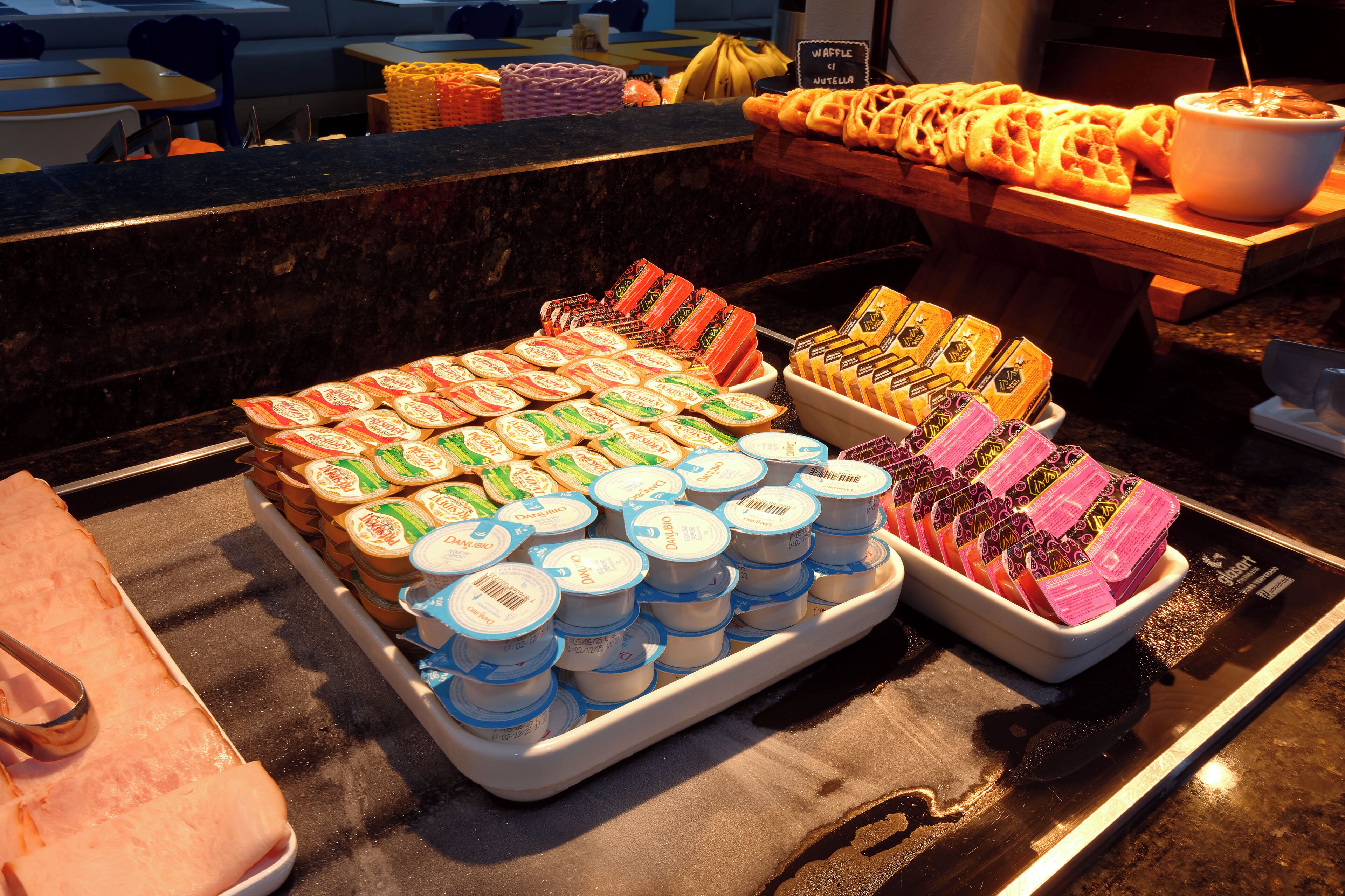 Buffet com diversos alimentos, incluindo waffles, doces e produtos embalados.