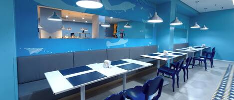 Un restaurante moderno con decoración azulada, que incluye una larga barra con mesas y sillas, y una zona de estar con bancos.