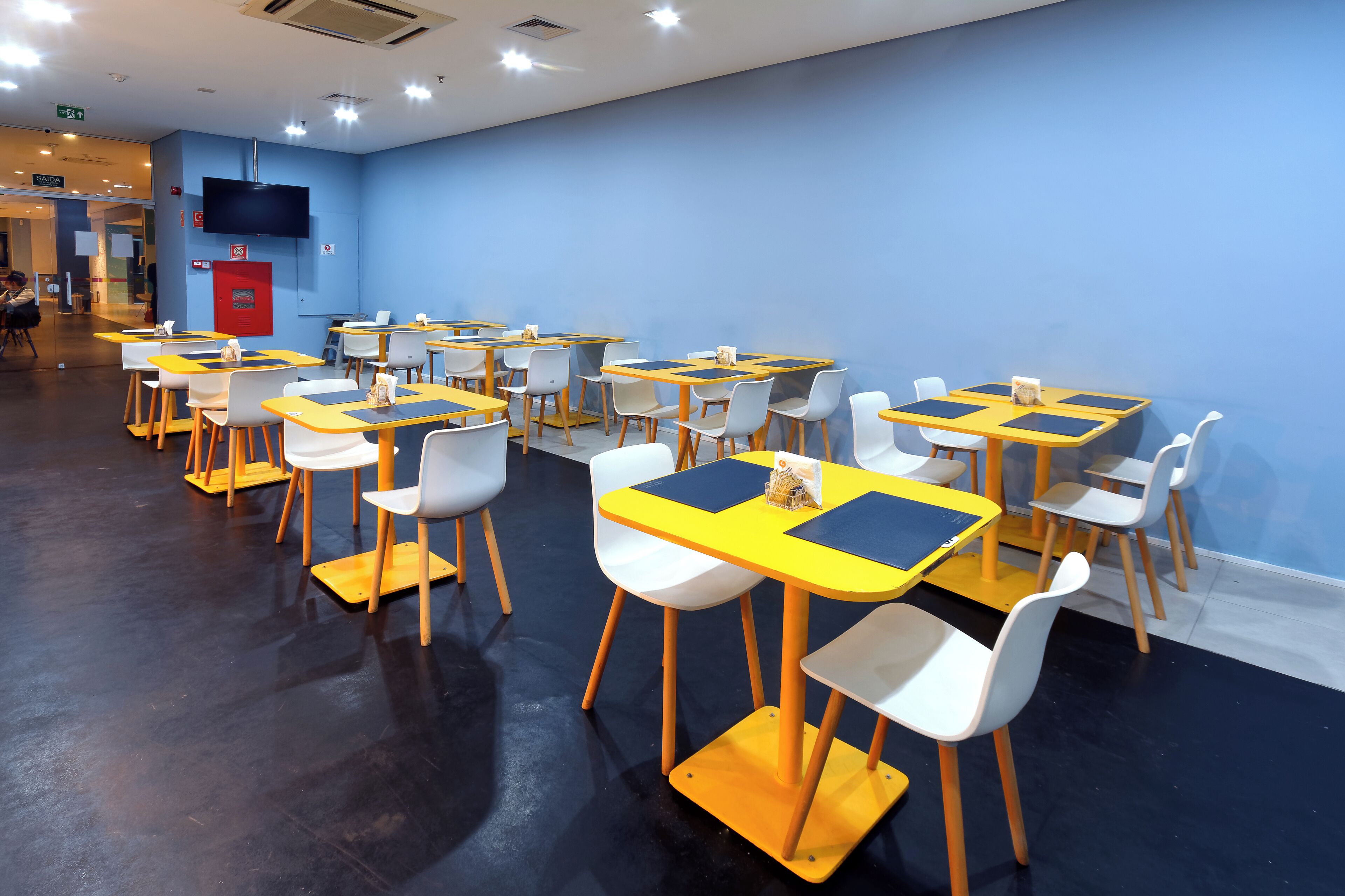 Un espace repas avec des tables et des chaises, un extincteur et un téléviseur fixé au mur.