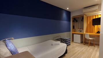 A hotel room with a bed, a desk, a chair, a small refrigerator, and an air conditioning unit.
