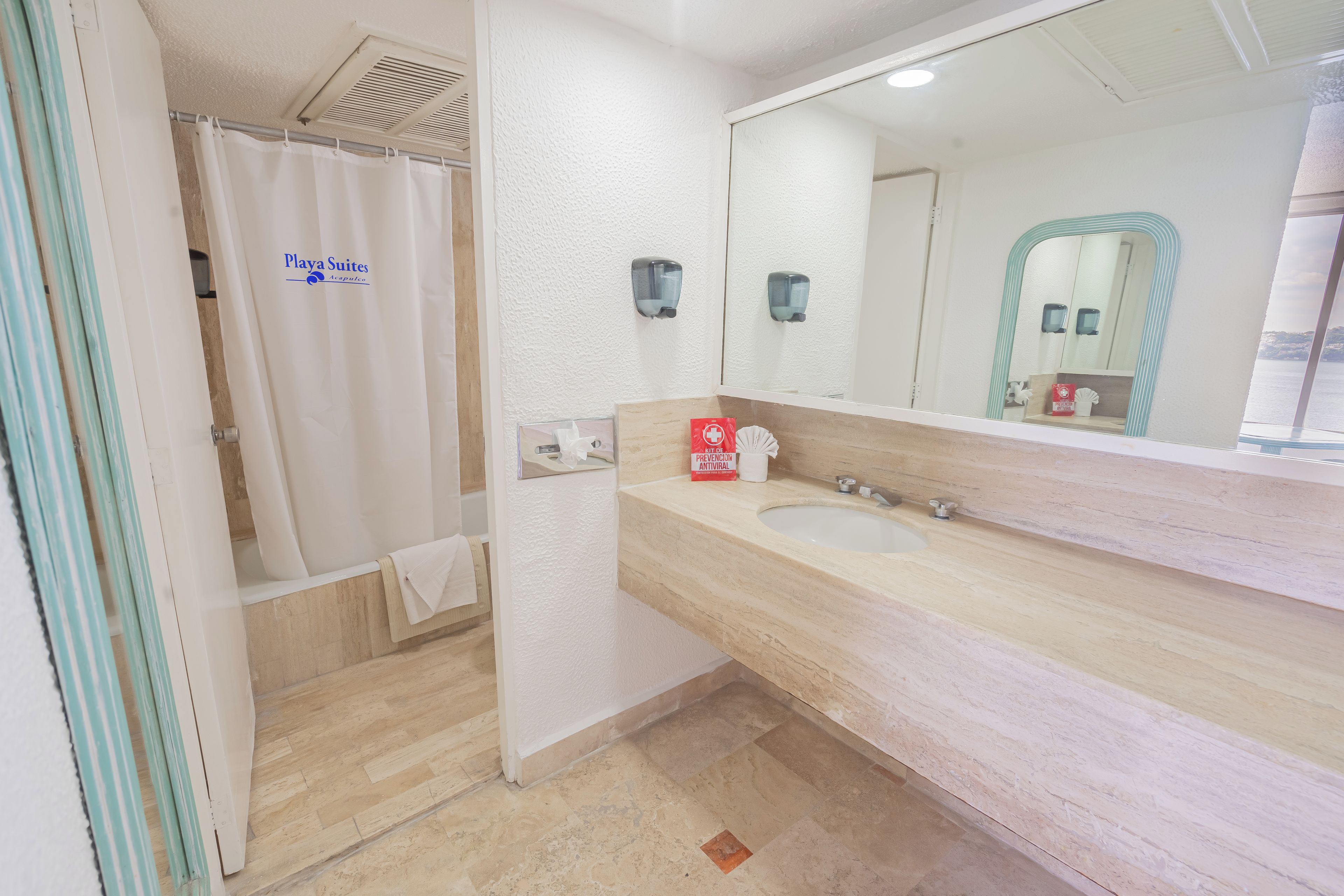 Un baño con cortina de ducha, lavamanos, espejo y dispensador de gel antibacterial.