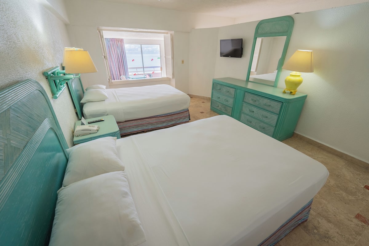Ein Schlafzimmer mit zwei Betten, einem Schminktisch, einem Spiegel, einem Fernseher und einem Fenster mit Vorhängen.