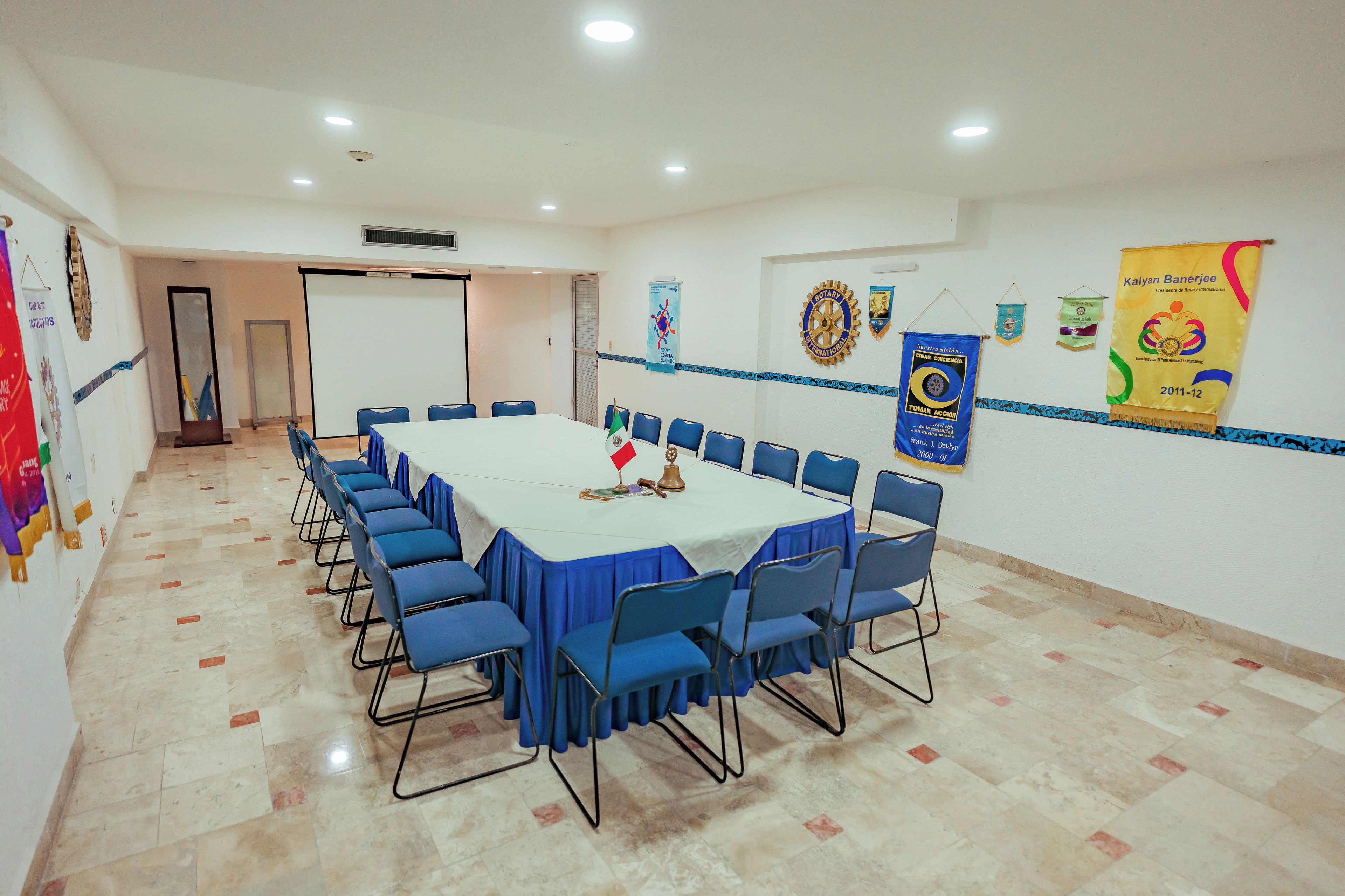 Un salón de conferencias con una larga mesa, sillas azules y un mantel blanco. Hay pancartas en las paredes y una puerta al fondo.