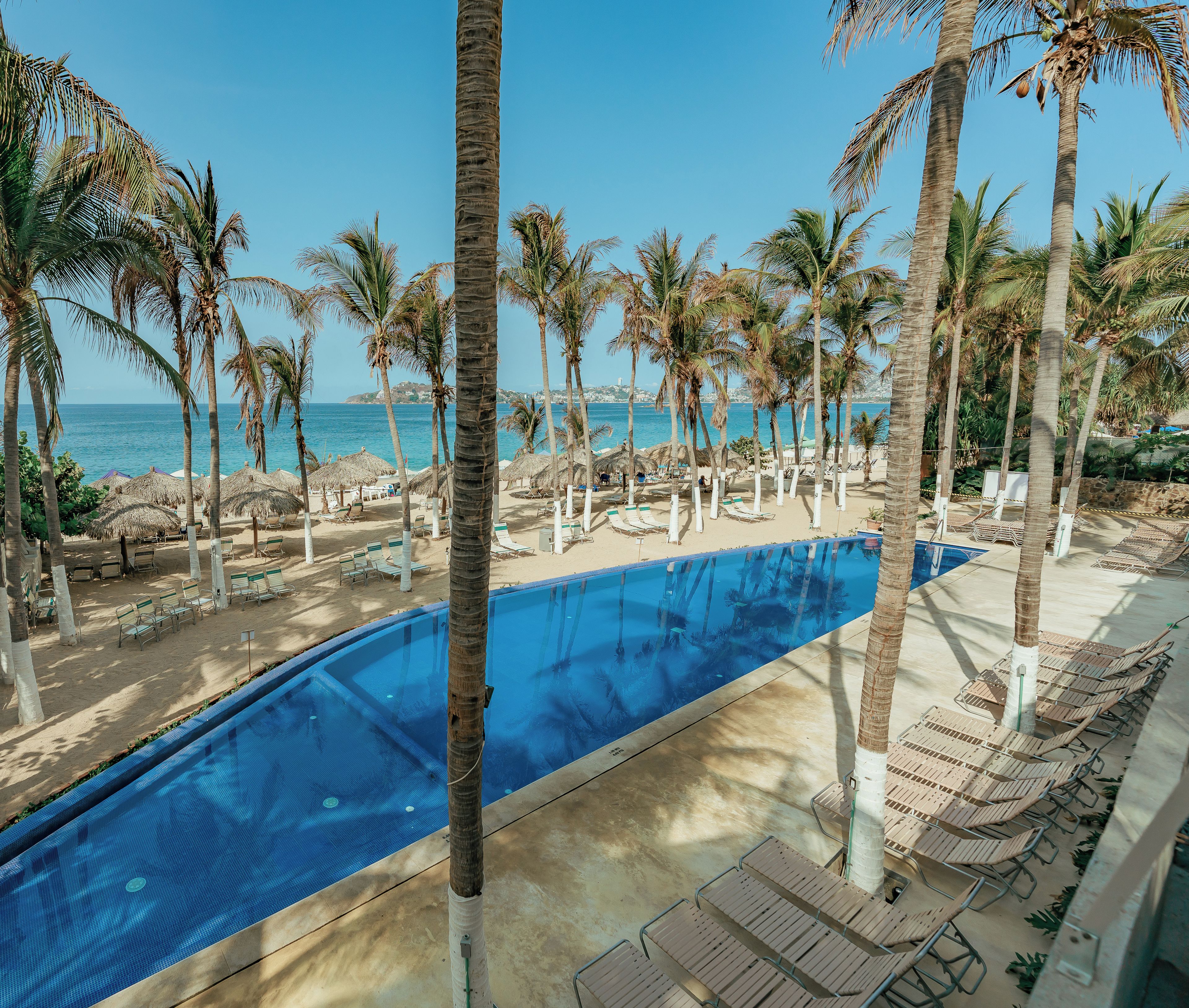 A poolside area with lounge chairs and palm trees, overlooking a beach and ocean.