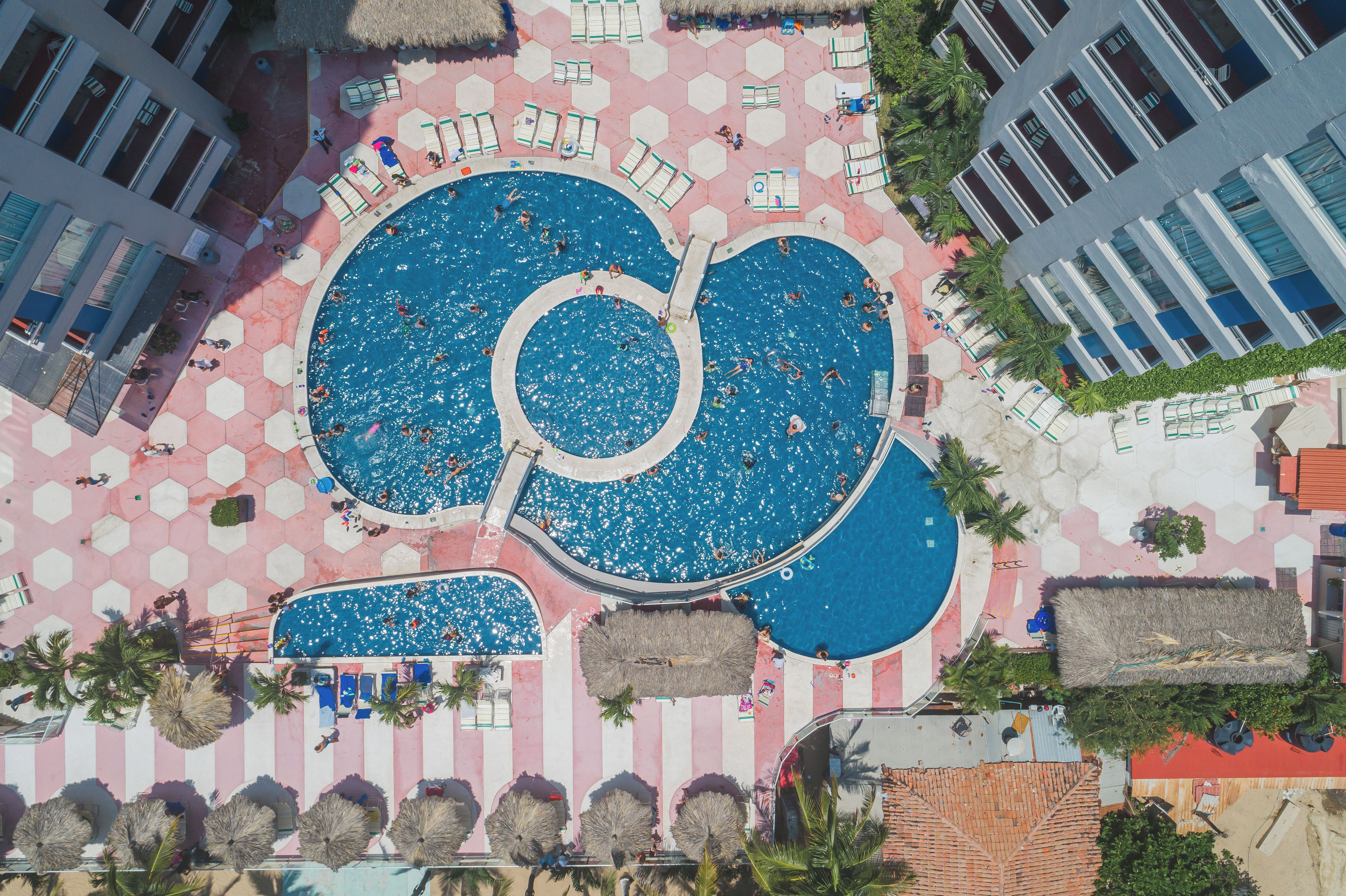 Vista aerea di un resort con diverse piscine, piastrelle esagonali e palme.