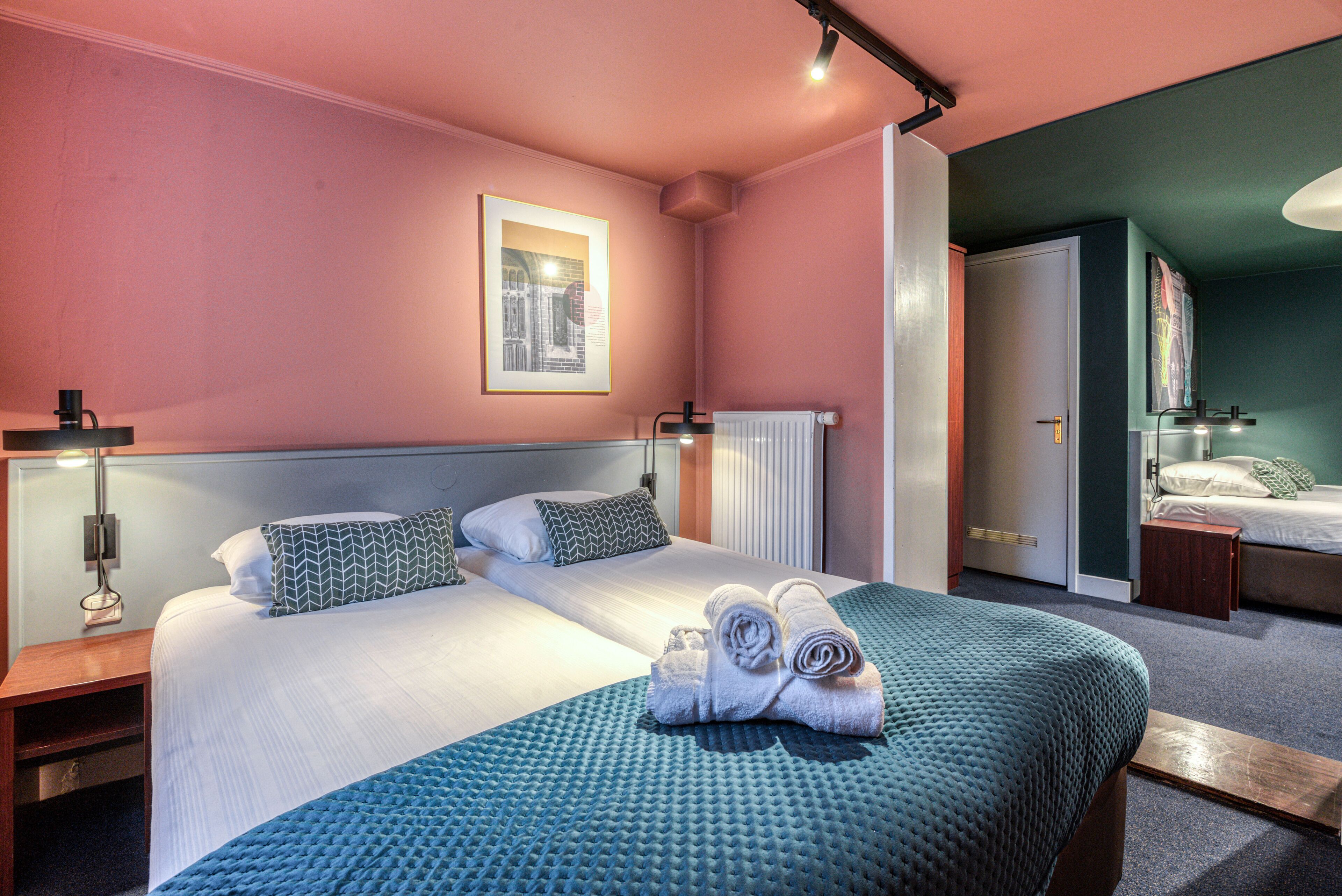 A hotel room with two beds, a large window, a radiator, and a bathroom visible through an open door.
