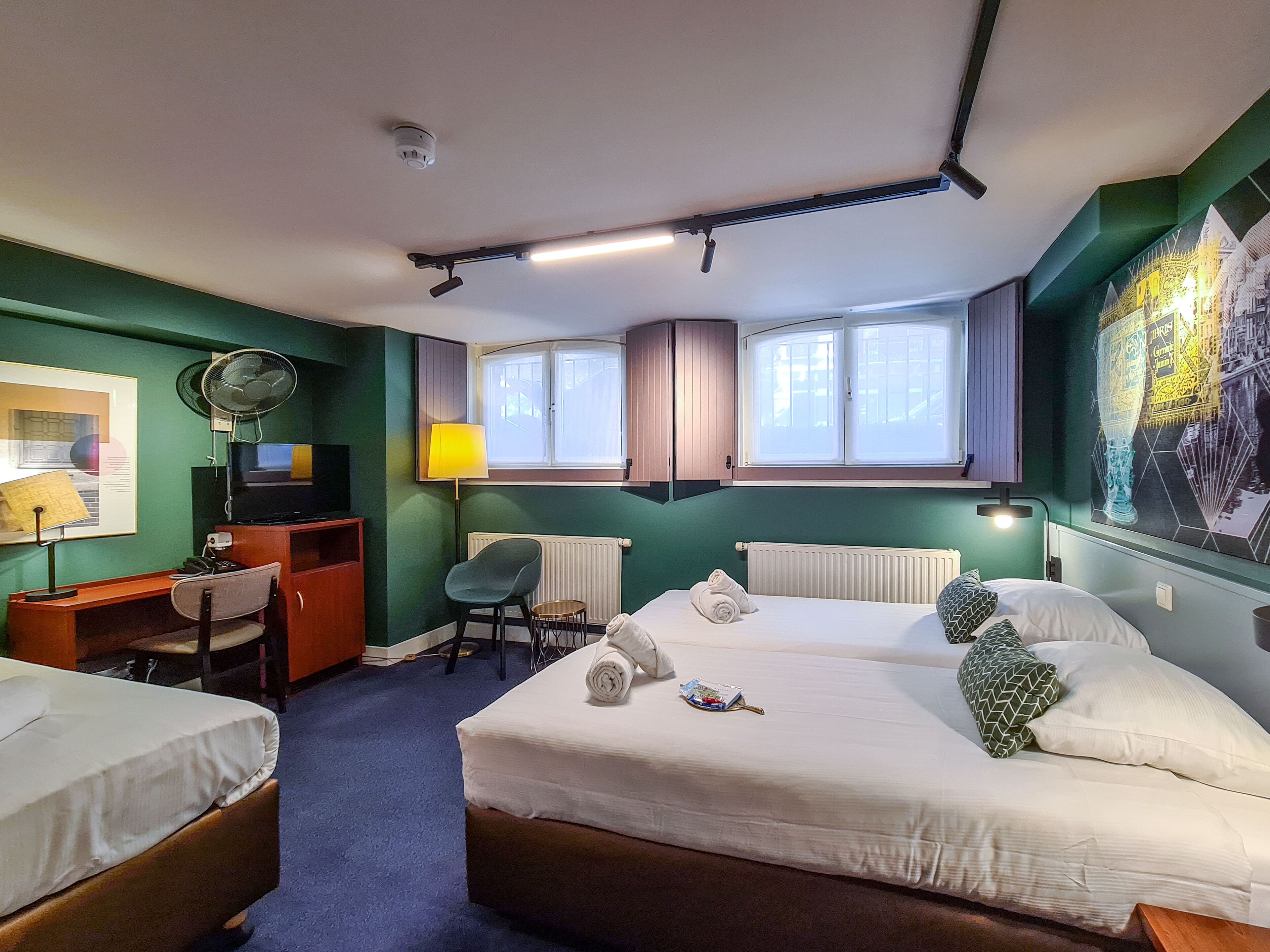 A hotel room with two beds, a desk, a chair, a radiator, and a window with a view.