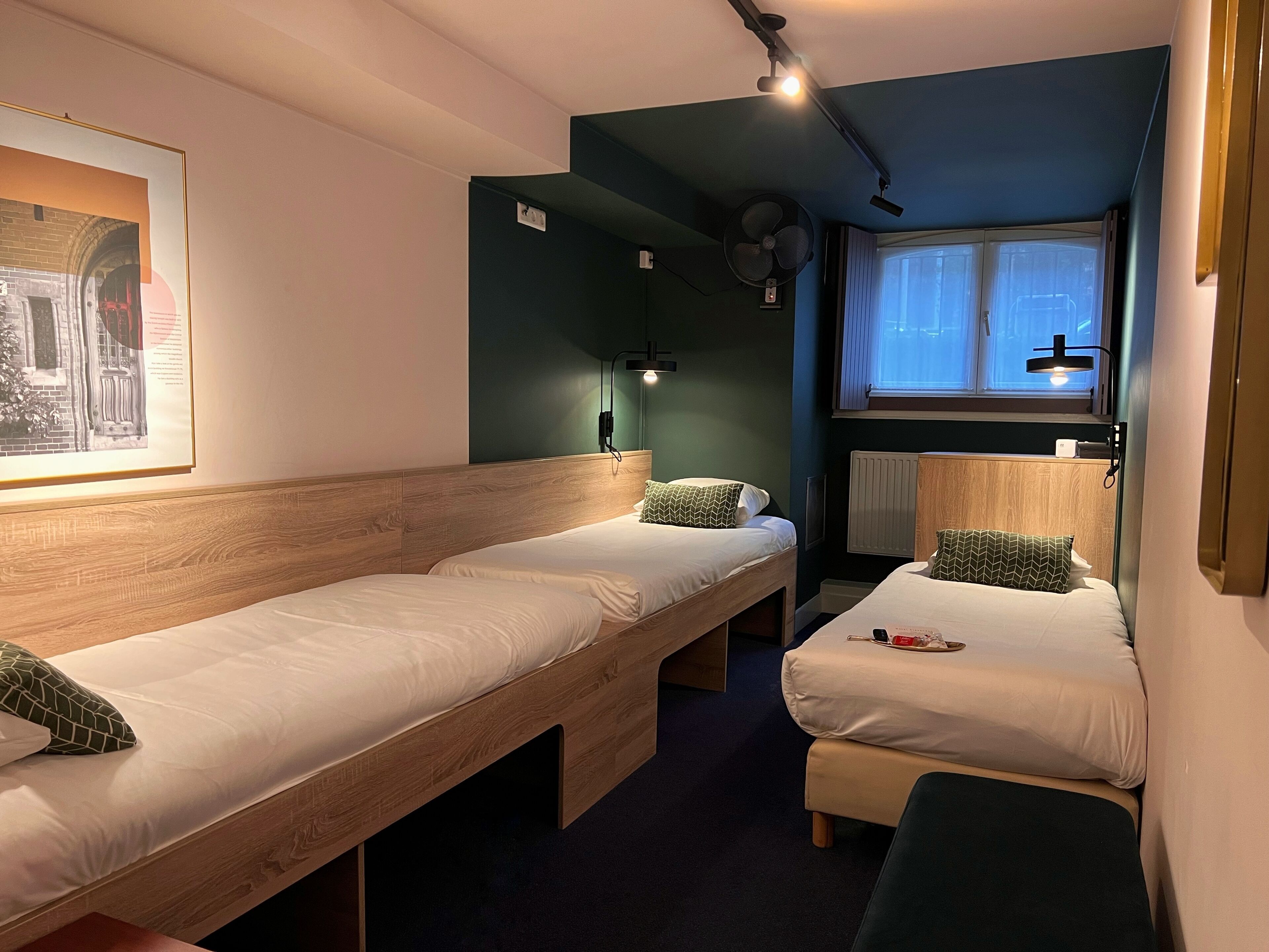 A room with two single beds, a bench, a wall-mounted fan, a window with blinds, and a framed picture on the wall.