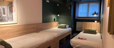 A room with two single beds, a bench, a wall-mounted fan, a window with blinds, and a framed picture on the wall.