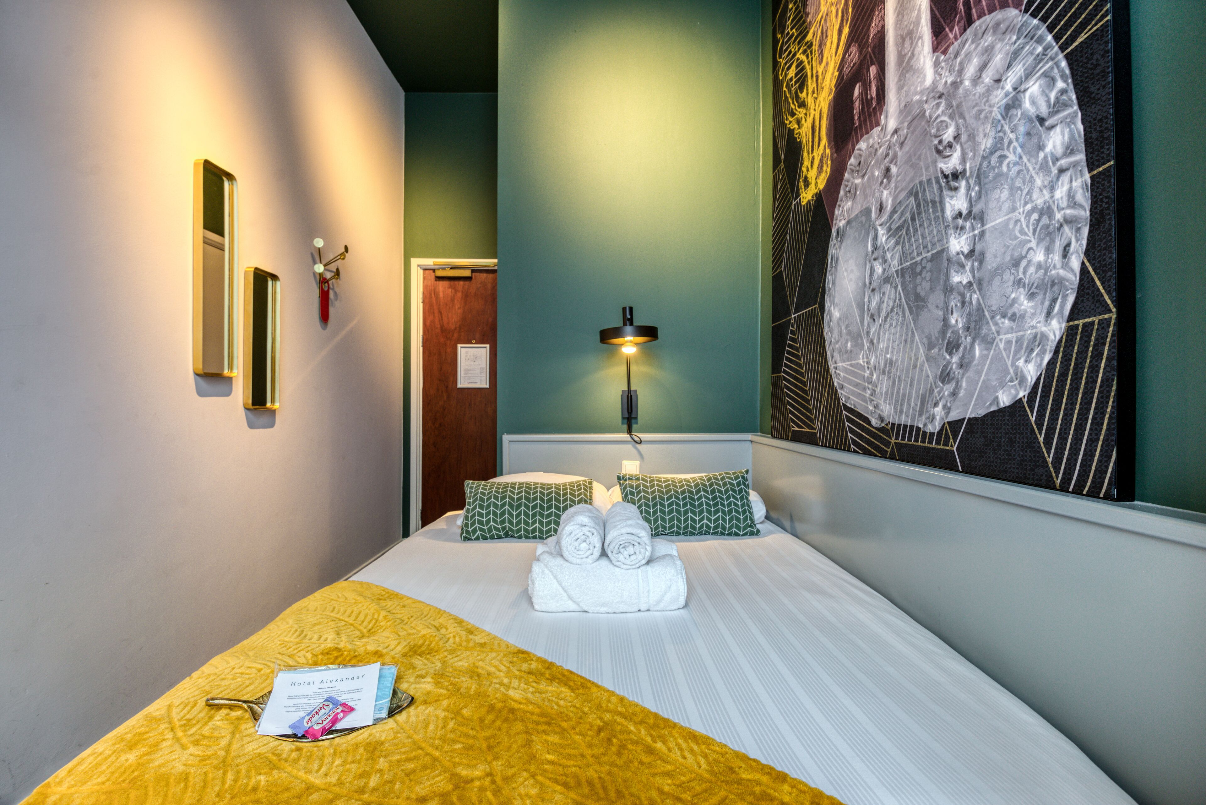 Une chambre d’hôtel avec un lit, une table de chevet, une lampe et un grand tableau abstrait accroché au mur.