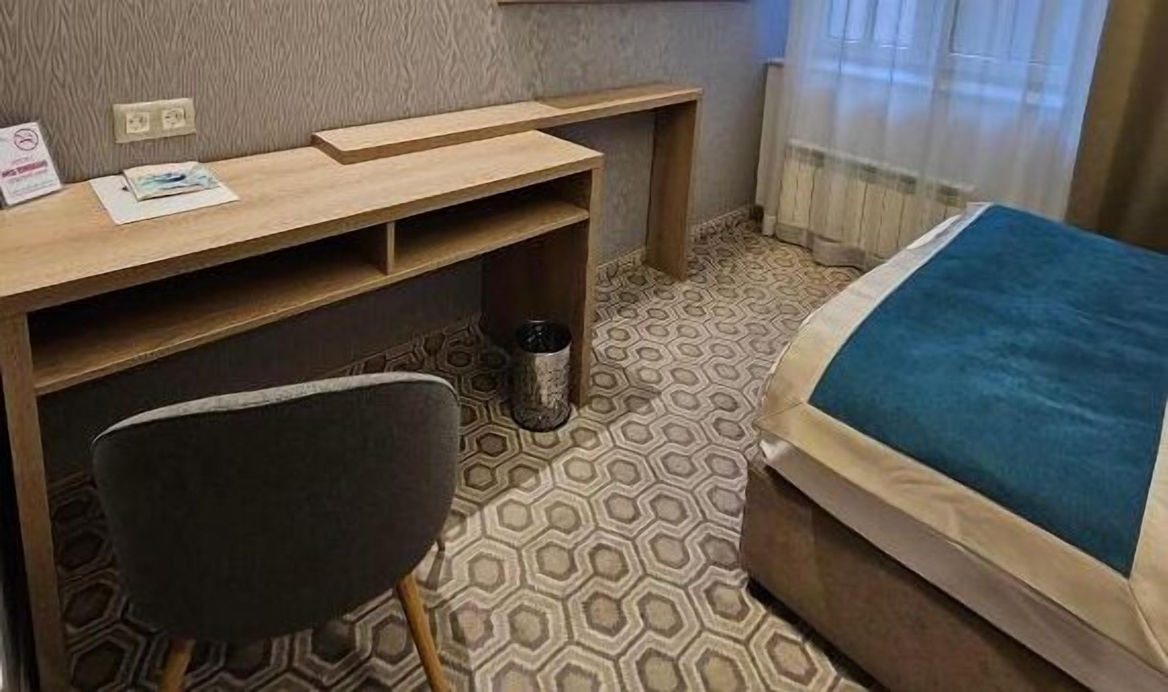 A hotel room with a wooden desk, a chair, a bed with a blue blanket, and a patterned floor.