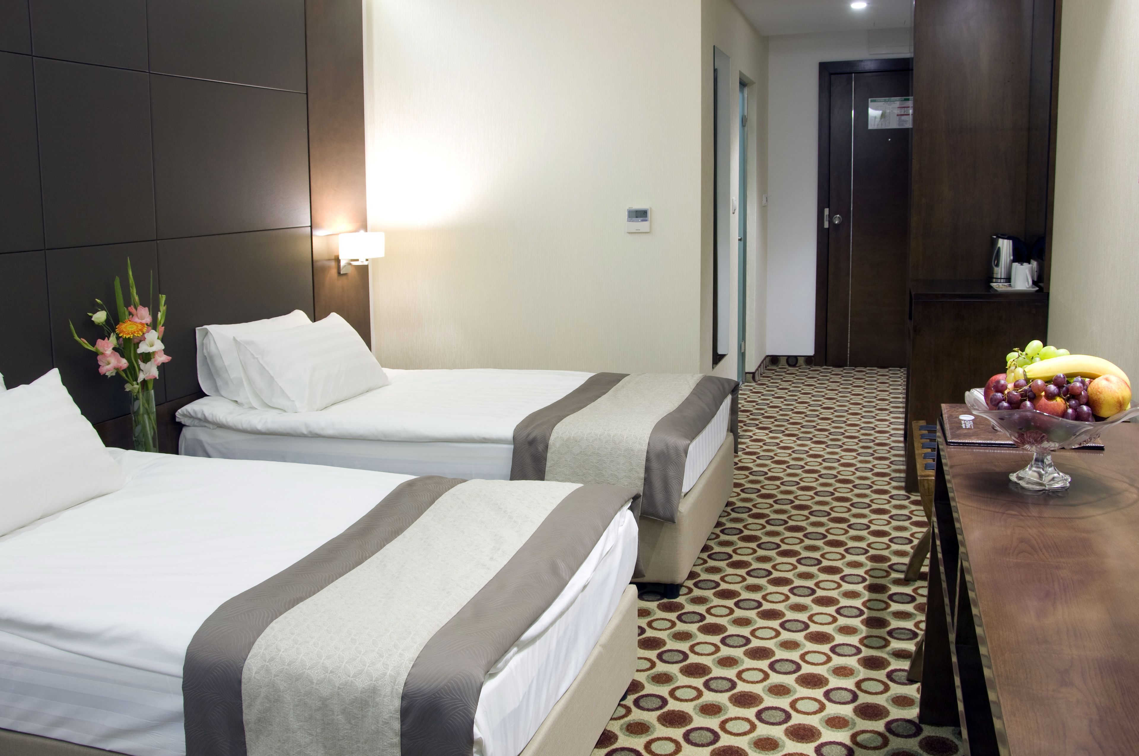 Habitación de hotel con dos camas, una mesita de noche con frutero, una lámpara de pared y una alfombra estampada.