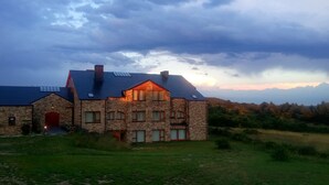 Una casa grande de piedra con múltiples ventanas y una puerta roja, rodeada de césped y colinas al fondo.
