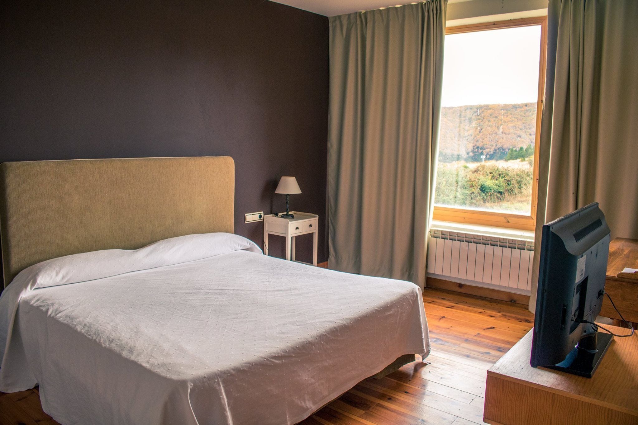 Un dormitorio con cama, mesita de noche, lámpara, televisor sobre un soporte de madera y ventana con cortinas.