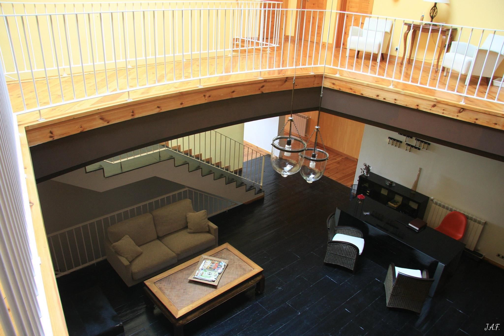 A spacious loft with a high ceiling, a staircase connecting two levels, a living area with a sofa and coffee table, and a kitchen area with a dining table and chairs.