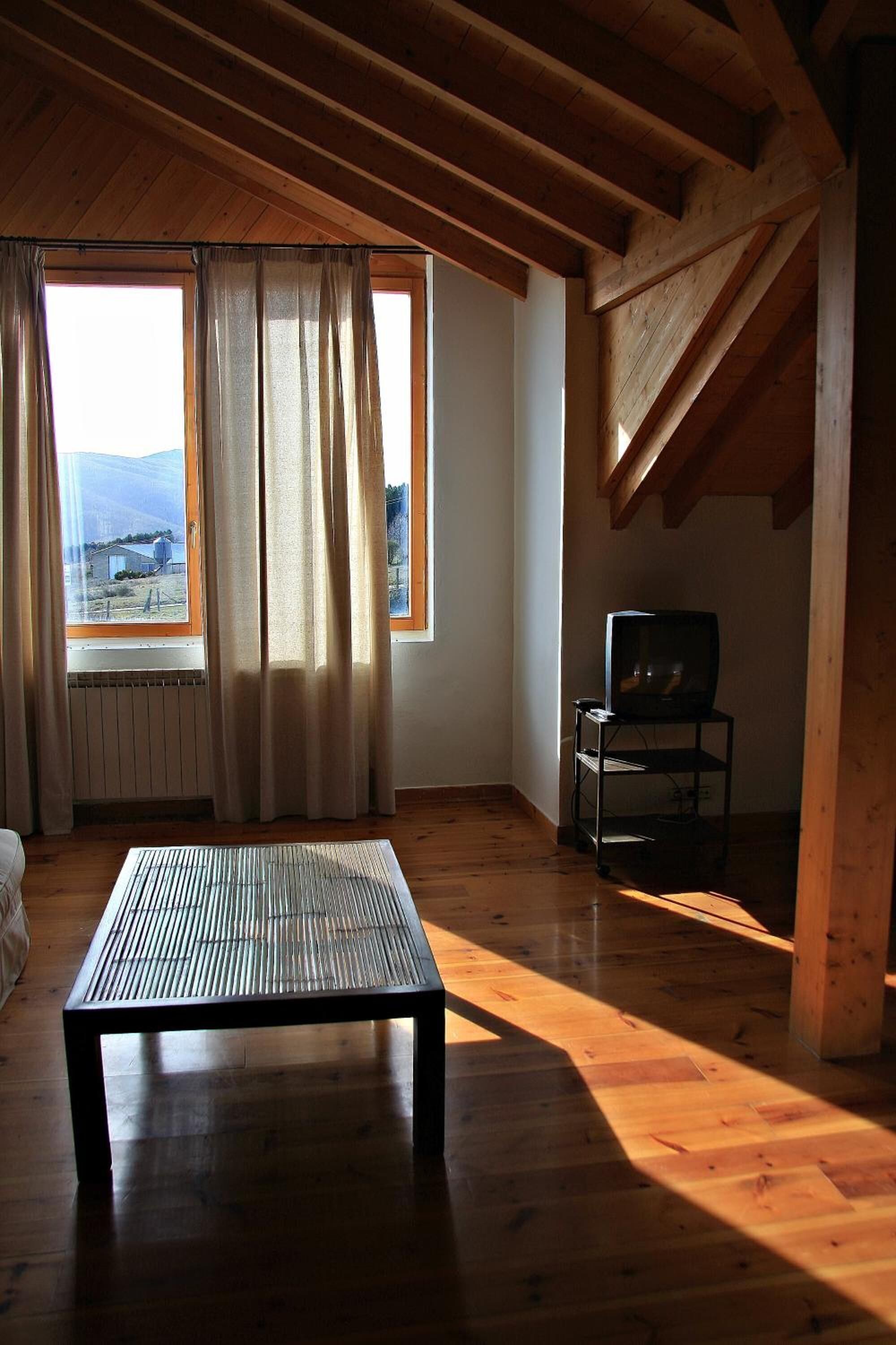 Une pièce avec un plancher en bois, une table, un téléviseur posé sur un meuble et une fenêtre avec des rideaux.