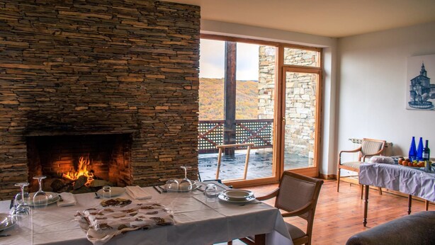 Un comedor con chimenea de piedra, una mesa dispuesta para una comida y vistas al exterior a través de grandes puertas correderas.