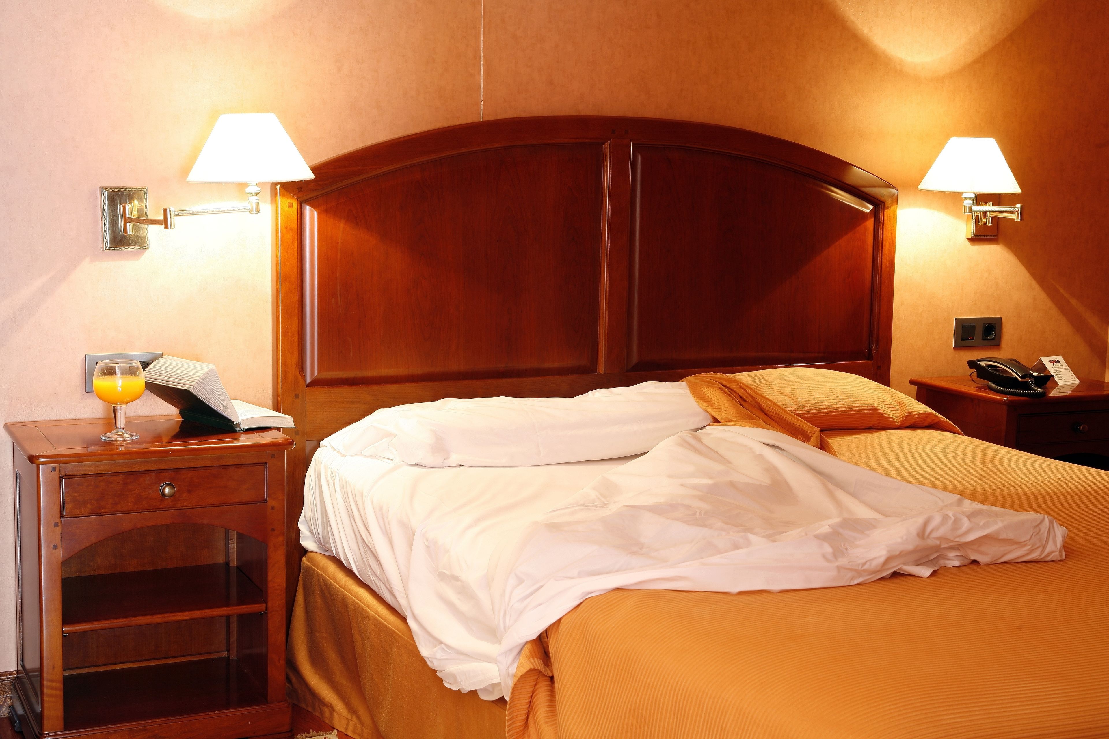 Habitación de hotel con cabecera de madera, dos lámparas de noche, una mesita de noche con teléfono, y otra mesita con un vaso de jugo de naranja y un libro.
