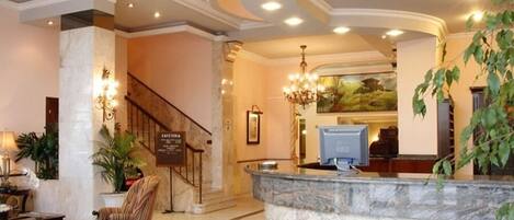 A hotel lobby with a marble reception desk, a staircase, and a chandelier.