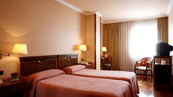 A hotel room with two beds, a desk, a chair, a television on a stand, and a window with curtains.