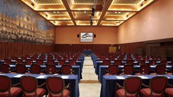 Un salón de conferencias amplio con filas de mesas y sillas, techo alto con luminarias, y un mural en la pared.