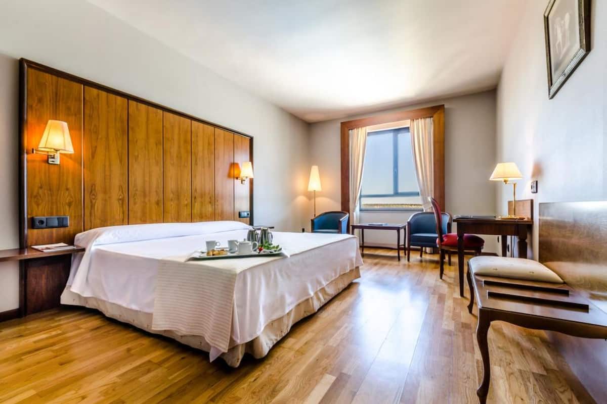 A hotel room with a large bed, a wooden headboard, two bedside lamps, a desk with a chair, and a window with curtains.