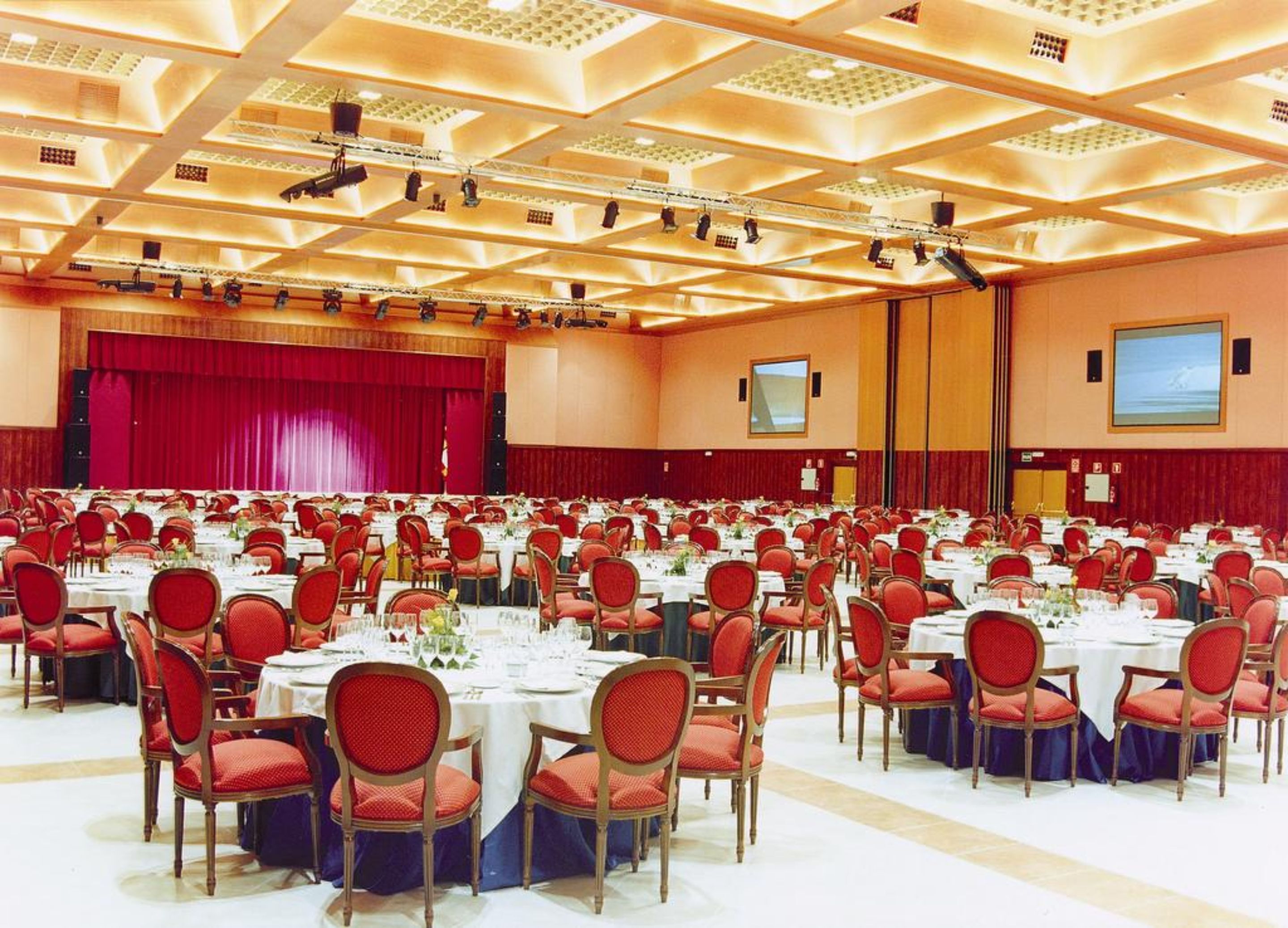 A large conference room with rows of tables and chairs, a stage with a red curtain, and a large screen on the wall.