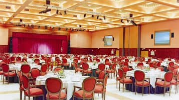 Un amplio salón de conferencias con filas de mesas y sillas, un escenario con cortina roja y una pantalla grande en la pared.