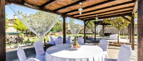 An outdoor patio with round tables and white chairs, a thatched roof, and a view of a garden and buildings in the background.