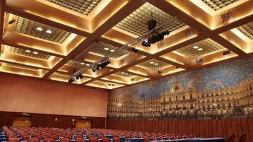 Un salón de conferencias con filas de mesas y sillas, un mural grande y un techo alto con un diseño de cuadrícula.