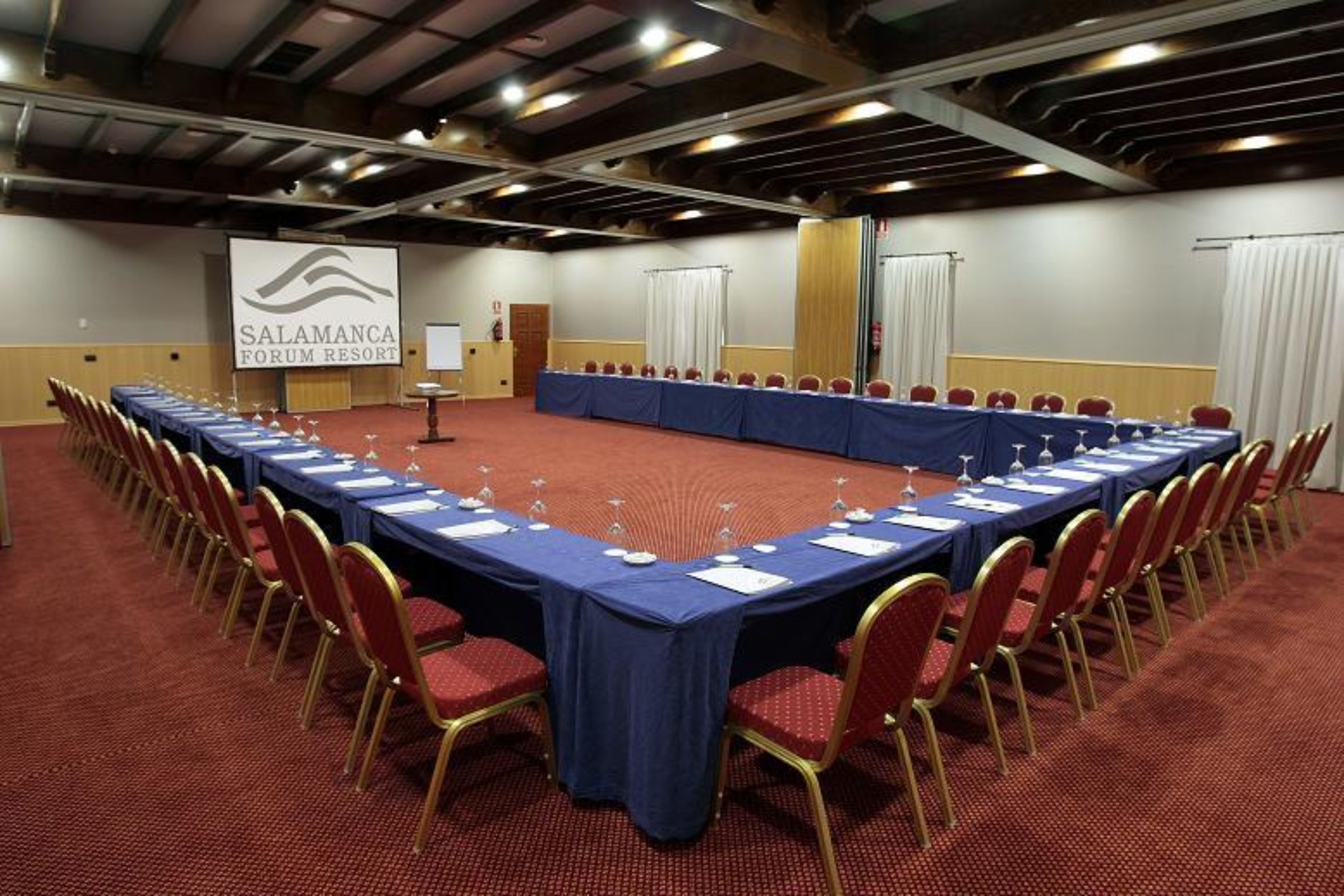 Ein Konferenzraum mit einem langen Tisch, roten Stühlen und einem Bildschirm, der 'SALAMANCA FORUM RESORT' anzeigt.