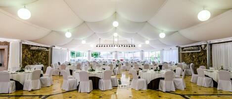 A banquet hall with round tables and chairs, a high ceiling with chandeliers, and a stone wall on one side.