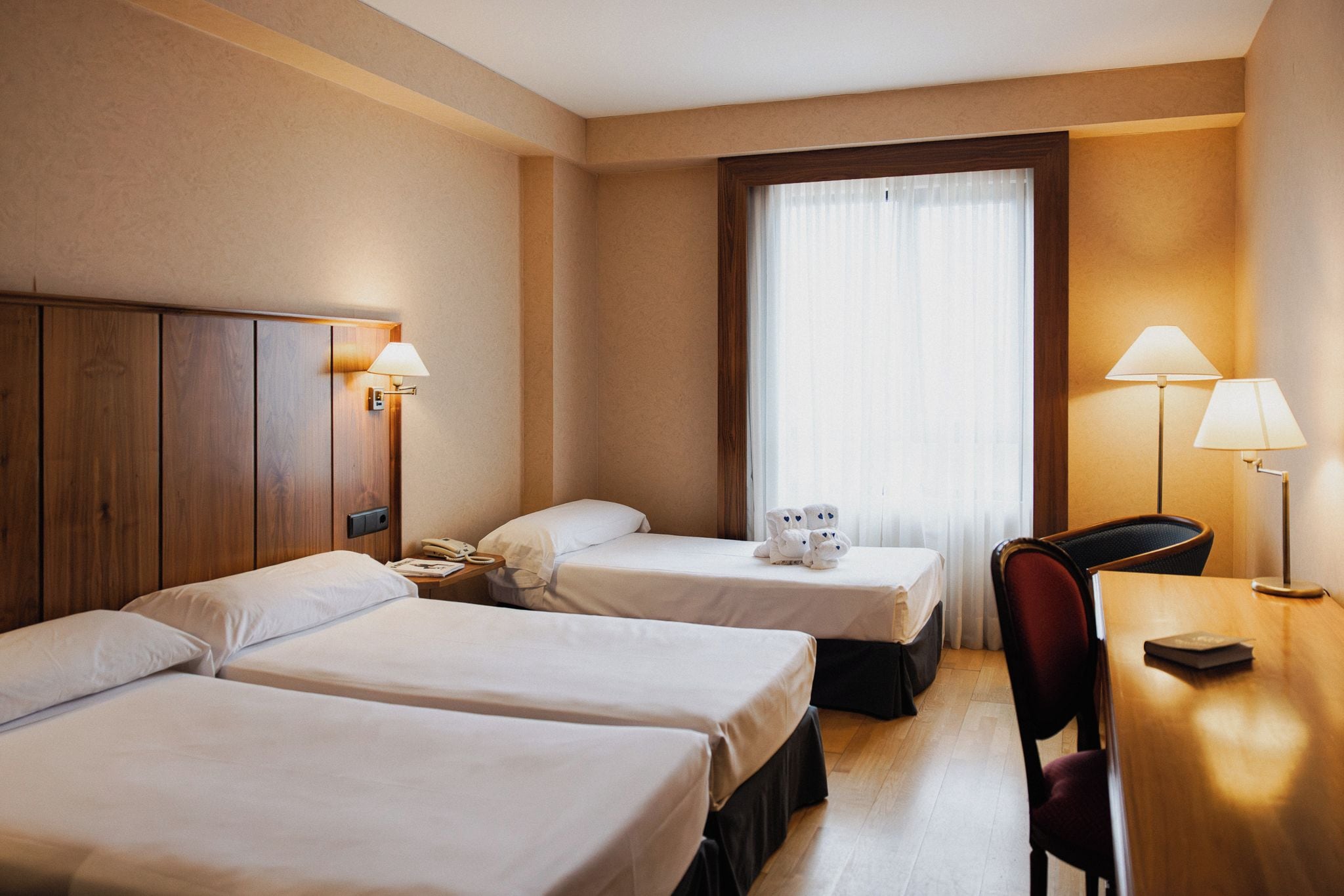 Ett hotellrum med två sängar, ett skrivbord, en stol, en lampa, en telefon och en nallebjörn.