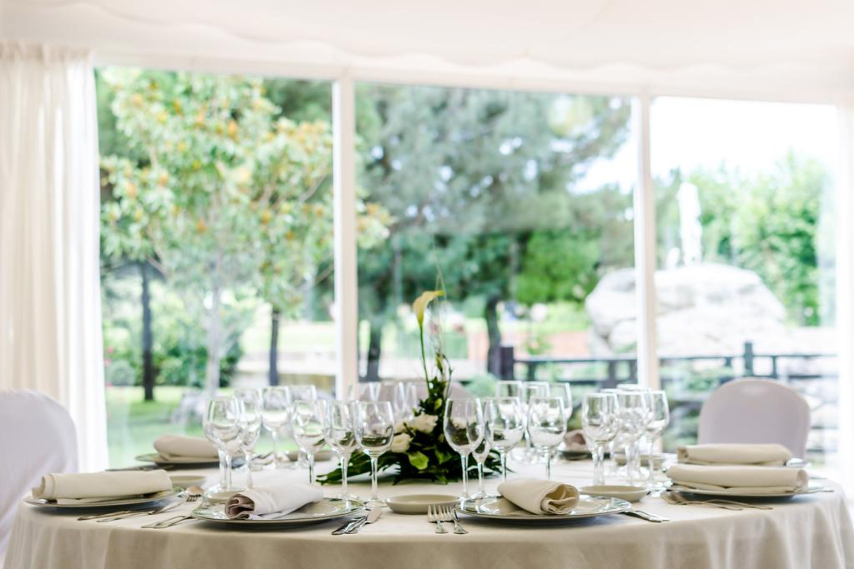 A table set for a formal dinner with white tablecloths, silverware, and wine glasses, set against a backdrop of greenery and a stone wall.