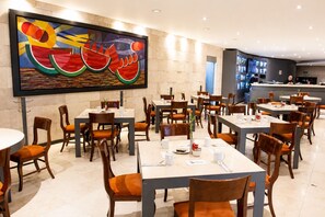 A restaurant with multiple tables and chairs, a large abstract painting on the wall, and a counter area in the background. - We Hotel Aeropuerto (Mexico City)