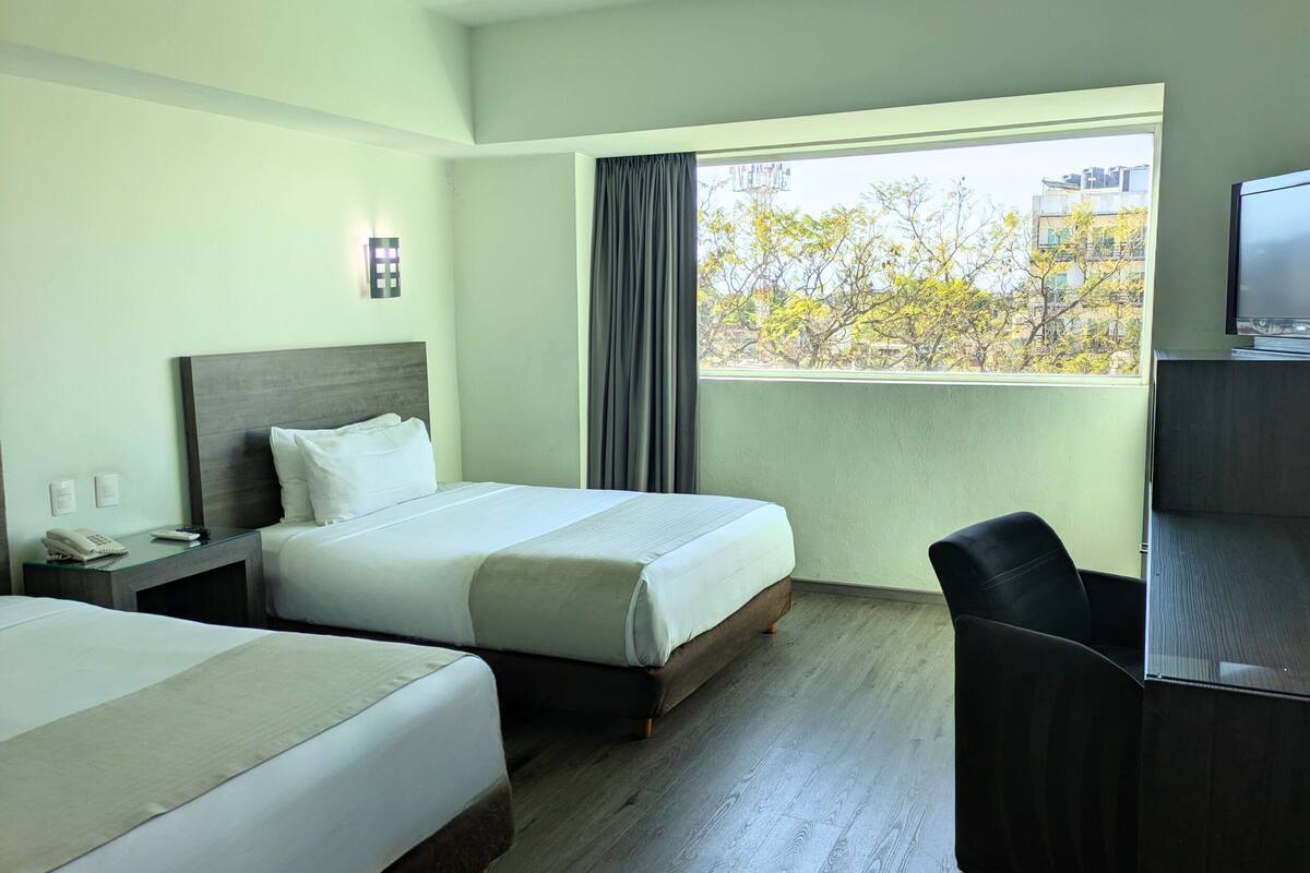Hotelkamer met twee bedden, een bureau met telefoon en afstandsbediening, een televisie en een raam met uitzicht op bomen en gebouwen.