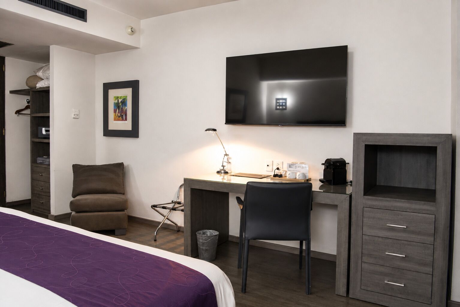 Habitación de hotel con cama, escritorio, silla, televisor, armario y cubo de basura.