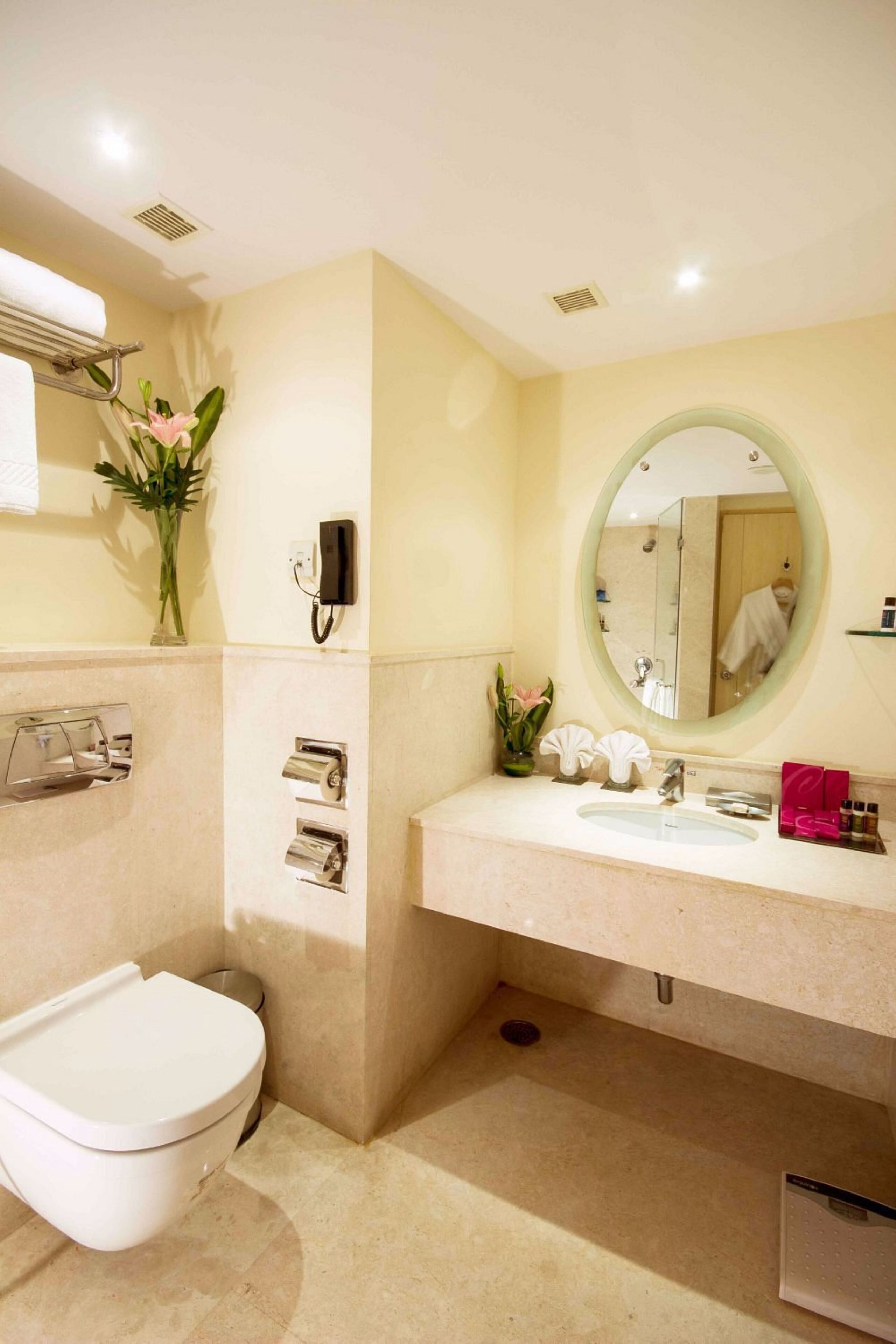 A bathroom with a toilet, sink, and mirror. There is a phone on the wall and a vase with flowers.