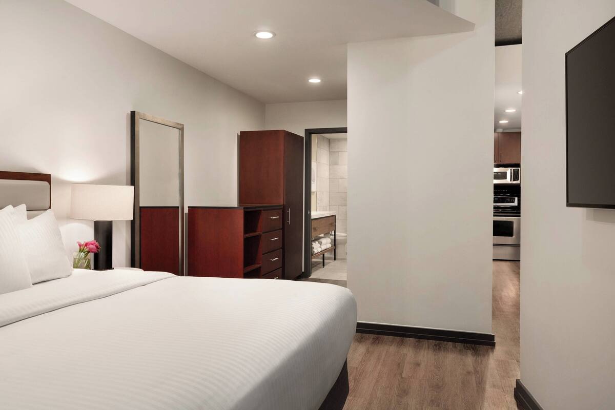 A hotel room with a large bed, a wooden wardrobe, a mirror, and a view into the kitchen area.