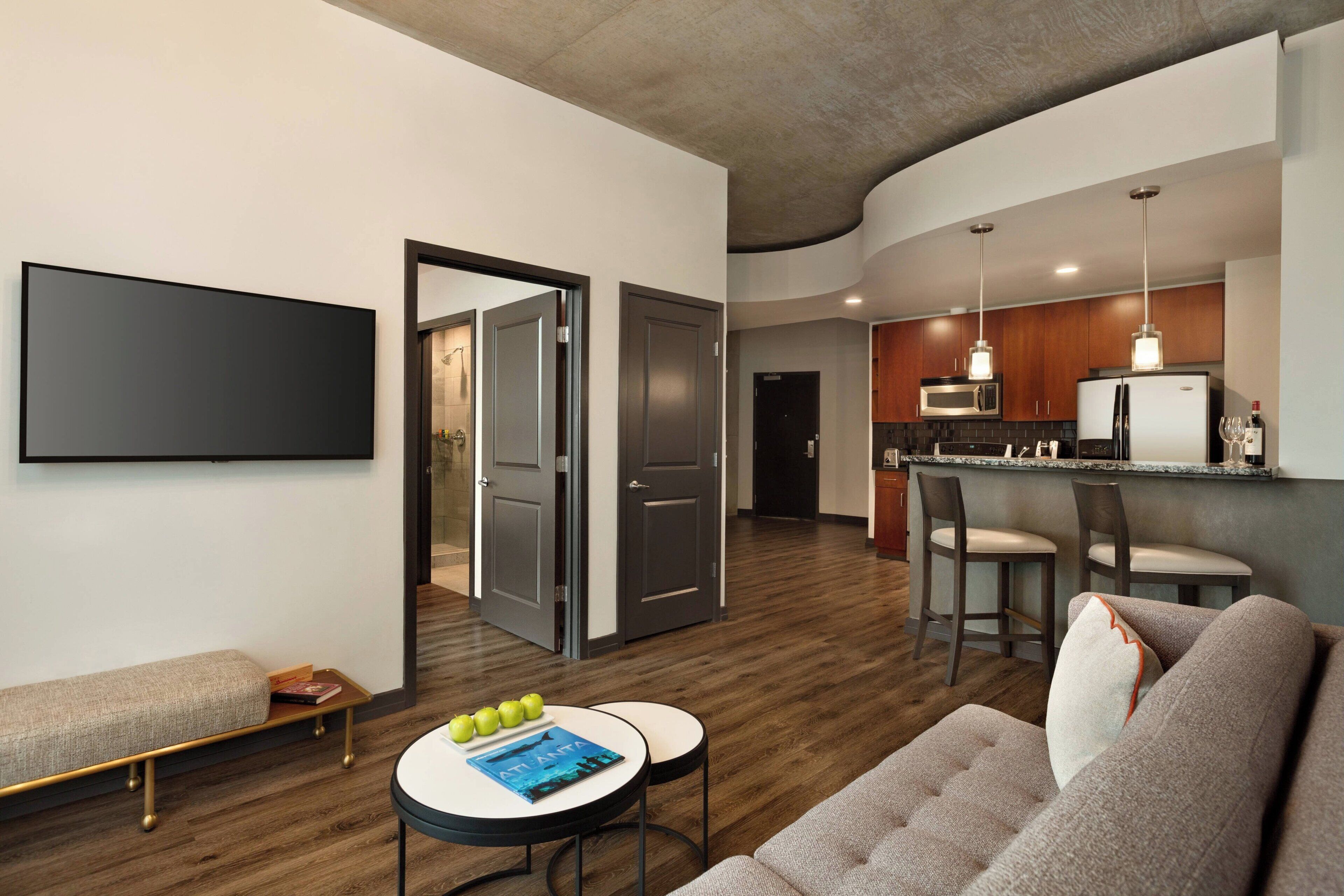 A modern living room with a sofa, a coffee table with books and apples, and a flat-screen TV mounted on the wall.