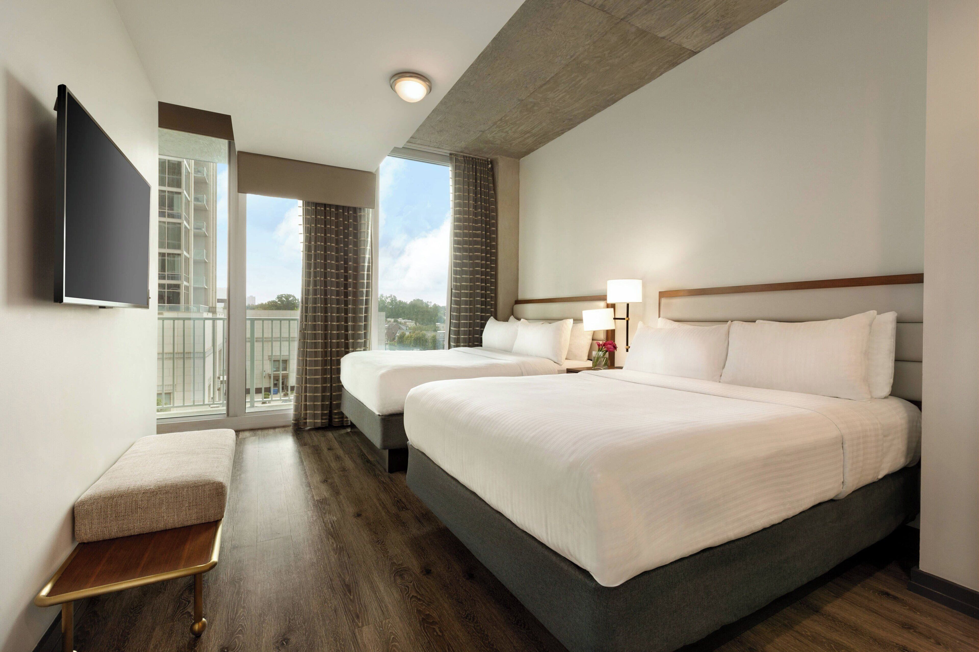 A hotel room with two beds, a flat-screen TV mounted on the wall, a small table, and a view of the outdoors through the window.