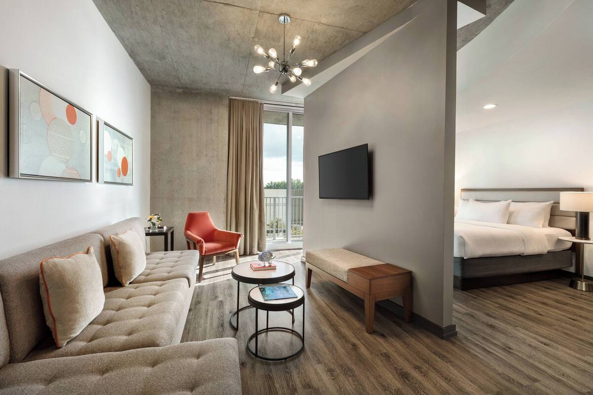 A modern hotel room with a sofa, a small round table, a bench, a bed, and a TV mounted on the wall.