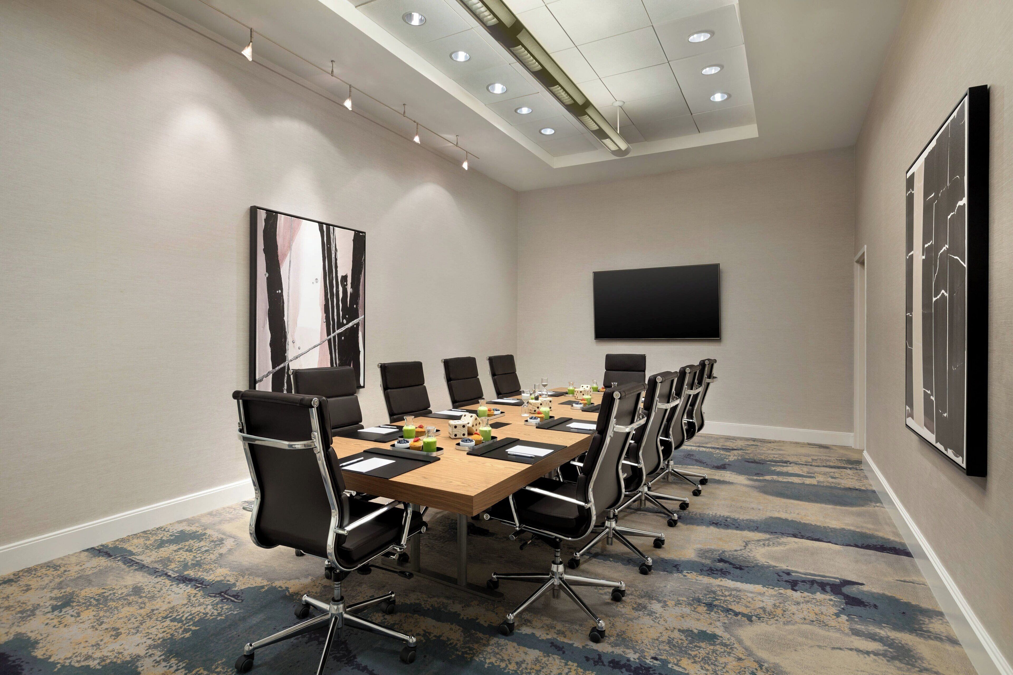 A conference room with a long table, black chairs, a flat-screen TV, and a patterned carpet.